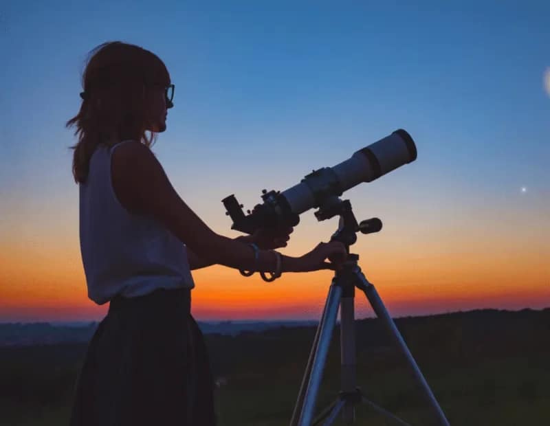 Mulher astrônoma com o seu telescópio ao fundo um céu com cores azul e alaranjado como se fosse o entardecer