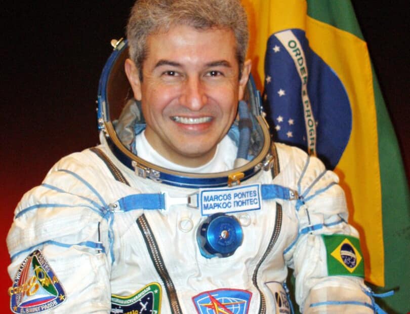 Marcos Pontes, astronauta brasileiro, sorri em sua foto oficial da NASA. Ele está usando um traje espacial branco, com detalhes em azul e vermelho, e não está usando o capacete. Seu traje possui a bandeira do Brasil no ombro esquerdo. Ao fundo, a bandeira do Brasil está visível, destacando a sua nacionalidade. A imagem transmite um ar de orgulho e conquista.