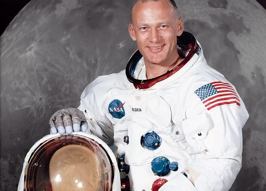 Foto oficial da NASA de Buzz Aldrin, o segundo homem a pisar na Lua durante a missão Apollo 11, sorrindo enquanto está de traje espacial branco, sem capacete. A imagem captura a Lua ao fundo, com suas mãos apoiadas no capacete, marcando um dos momentos mais icônicos da história da exploração espacial.