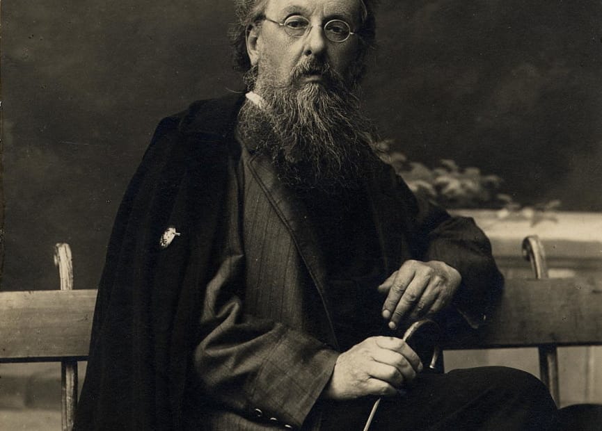 Konstantin Tsiolkovski, um homem idoso com uma longa barba branca e óculos redondos, está sentado em um banco. Ele segura uma bengala com as mãos, transmitindo uma postura serena e contemplativa. Considerado um dos pioneiros da astronáutica, suas ideias ajudaram a moldar o futuro das viagens espaciais.