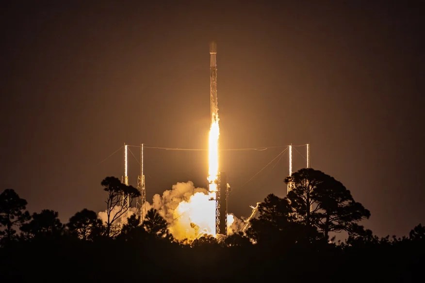 O foguete Falcon 9 da SpaceX sendo lançado da plataforma de decolagem, iluminando o céu com sua poderosa queima de motores. O evento ocorreu em 17 de fevereiro de 2025, marcando mais um avanço na reutilização de foguetes para missões espaciais. No fundo, pode-se ver a estrutura da plataforma de lançamento e uma leve camada de nuvens, criando um contraste impressionante com a luminosidade do foguete em ascensão.