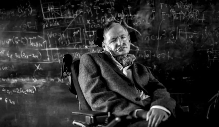 Stephen Hawking de frente, com um quadro negro ao fundo repleto de cálculos e fórmulas matemáticas escritas em giz branco. A imagem transmite um ambiente de estudo e reflexão científica.