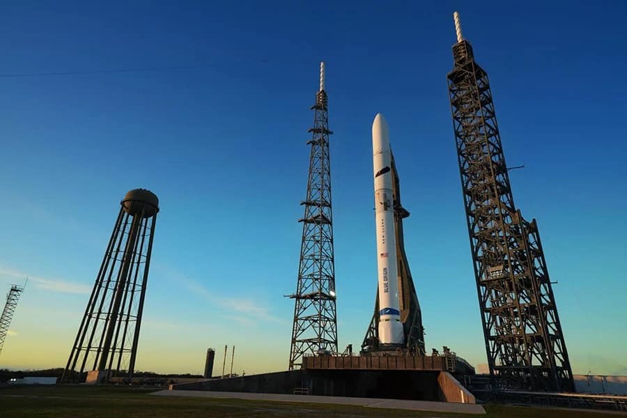 O foguete New Glenn da Blue Origin, de cor branca, está posicionado na plataforma de lançamento durante o dia, aguardando o momento para iniciar seu voo. O céu azul ao fundo contrasta com a estrutura imponente do foguete, pronto para sua missão.