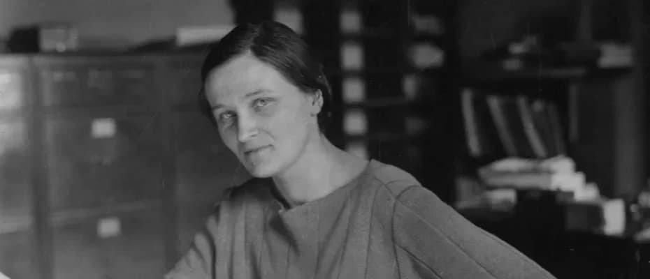 Cecilia Payne-Gaposchkin sentada em uma cadeira, apoiada em uma mesa de escritório, usando roupas da época. A cena é ambientada em uma sala de aparência clássica, com móveis antigos, refletindo o contexto histórico de sua época.
