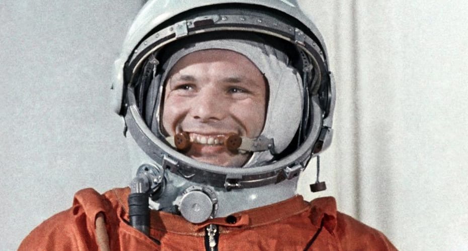 Yuri Gagarin, vestido com o traje espacial soviético laranja e um capacete branco com as letras "CCCP" na frente, exibe um sorriso radiante. Seu olhar transmite confiança e entusiasmo, refletindo o espírito pioneiro da primeira viagem tripulada ao espaço.