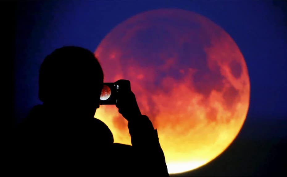 Silhueta de uma pessoa tirando uma foto da Lua de Sangue. A figura humana está posicionada de lado, com um celular em mãos, focando na Lua vermelha no céu noturno. O contraste entre a silhueta escura e o brilho avermelhado da Lua cria um efeito dramático e misterioso, destacando a raridade do evento celestial.