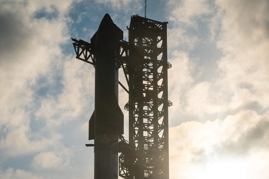 Imagem da Starship da SpaceX antes do sexto teste de voo, posicionada na plataforma de lançamento. O foguete está montado em sua estrutura de lançamento, com os motores e sistemas de propulsão visíveis. O céu claro ou parcialmente nublado no fundo destaca a imponente presença do foguete, que se prepara para sua missão espacial. Detalhes dos suportes e da infraestrutura de lançamento complementam a cena, mostrando a preparação meticulosa para o voo