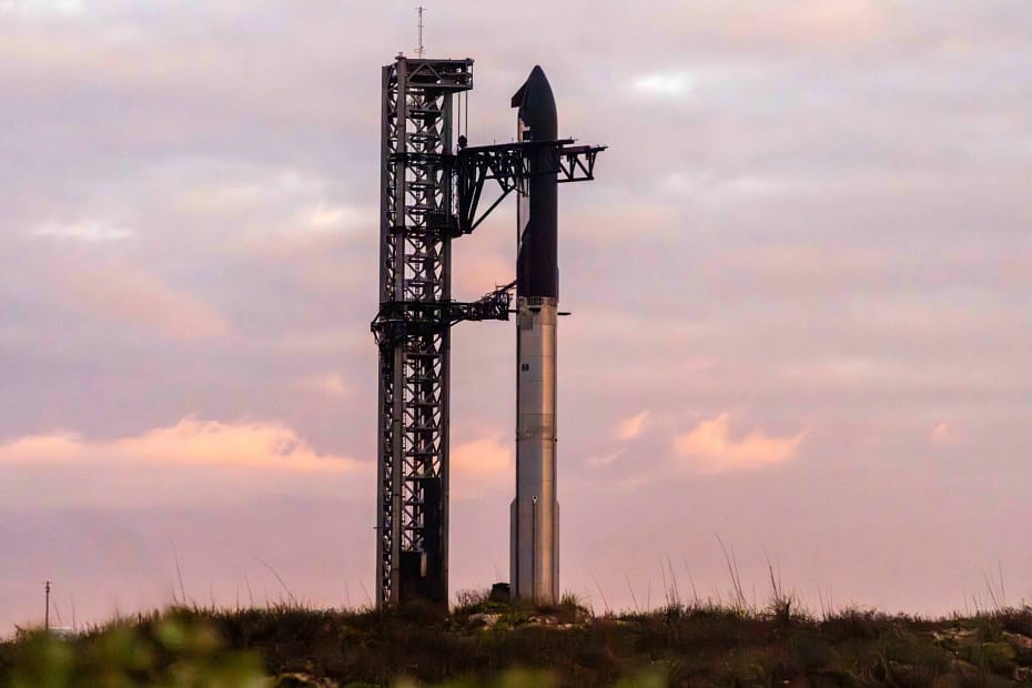 Foguete Starship aguardando o lançamento, com o booster metálico visível e a nave Starship em cor preta. O foguete está posicionado na plataforma de lançamento, enquanto o céu ao fundo adiciona um contraste suave à cena.