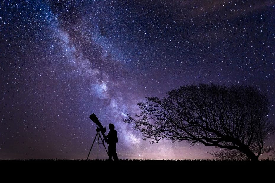 Silhueta de uma pessoa com um telescópio apontado para o céu estrelado, com cores roxas e lilás representando uma galáxia distante.