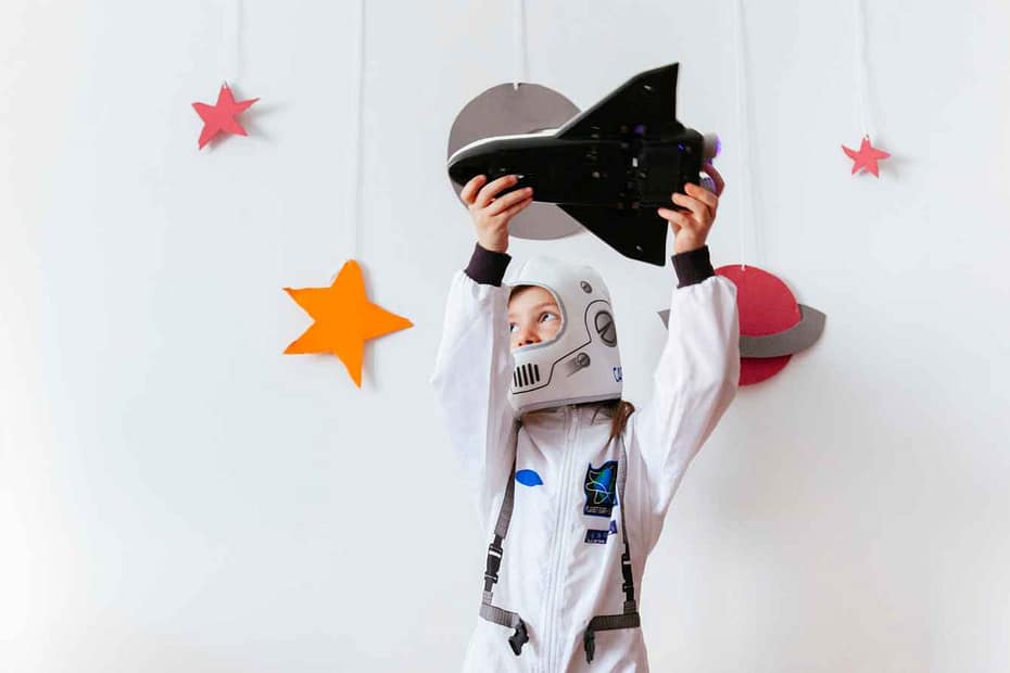 Criança com traje de astronauta branco brincando com um foguete de brinquedo, com recortes de planetas e estrelas na parede branca ao fundo.