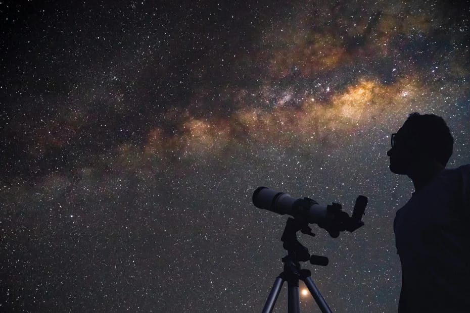 Astrônomo com telescópio apontado para o céu noturno estrelado. A imagem ilustra como observar o céu e apreciar fenômenos como chuvas de meteoros e estrelas cadentes.