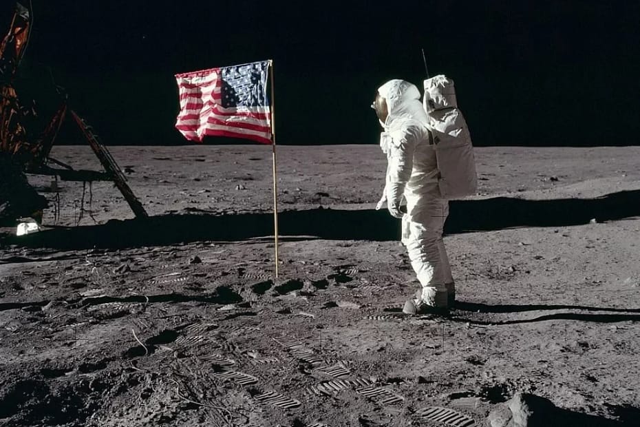 Apollo 11 Buzz Aldrin, o astronauta da missão Apollo 11, saudando a bandeira dos Estados Unidos na superfície da Lua em 1969. Ele está vestido com seu traje espacial, com o visor refletindo a paisagem lunar ao fundo, enquanto a bandeira americana ondula ao vento (apesar da falta de atmosfera) em um gesto simbólico de conquista.