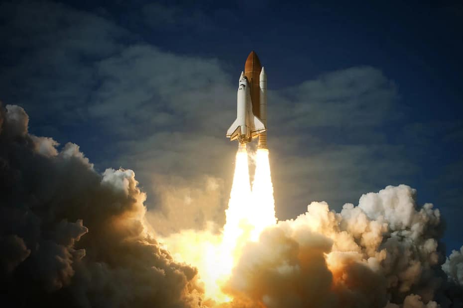 Lançamento de um ônibus espacial, com a enorme potência dos foguetes gerando uma explosão de fogo e fumaça enquanto o veículo ascende em direção ao céu. O ônibus espacial é visto no início de sua trajetória, com a separação dos boosters laterais e o veículo se afastando da base de lançamento. A palavra-chave "Lançamento espacial" é representada pelo evento dinâmico do lançamento.