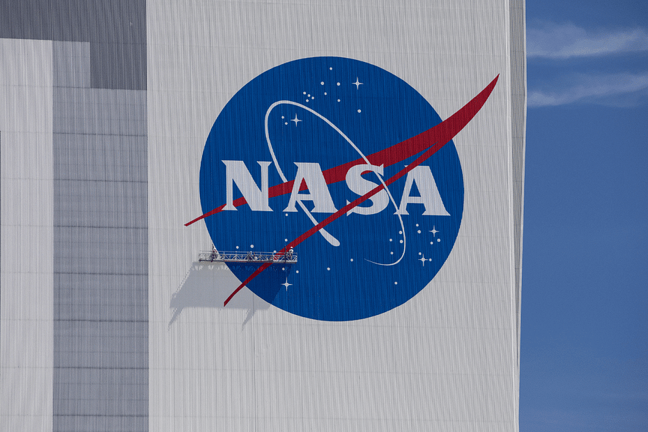 Imagem do logo da NASA na fachada do Kennedy Space Center, com trabalhadores realizando manutenção na área. O logo, de grande escala, ocupa grande parte da fachada, e os trabalhadores, em tamanho reduzido devido à magnitude do logo, parecem minúsculos em comparação com a imponente estrutura. A cena transmite a grandiosidade do local e a dedicação dos profissionais envolvidos na conservação do símbolo.