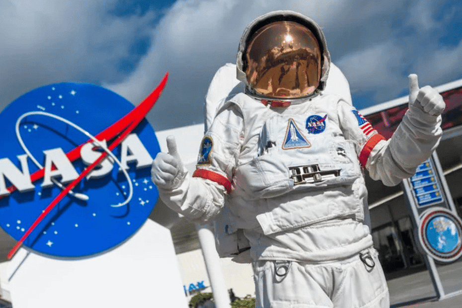 ecepção do Kennedy Space Center, com o logo grande da NASA ao fundo. Em primeiro plano, um astronauta usando traje espacial faz o sinal de 'joia' com as mãos, convidando os visitantes a conhecerem mais sobre o que é a NASA, onde fica e como funciona