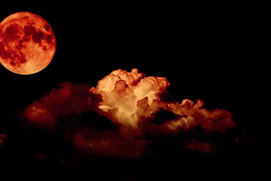 eclipse lunar total, conhecido como Lua de Sangue. A Lua aparece à direita da imagem, com uma tonalidade avermelhada intensa. Do lado esquerdo, grandes nuvens ocupam parte do céu, refletindo algumas tonalidades vermelhas devido à luz dispersa na atmosfera.