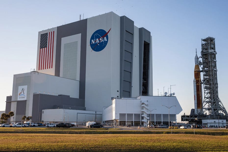 Vista distante do Kennedy Space Center ao amanhecer, com o foguete SLS (Space Launch System) no pad de lançamento. Ao lado, a bandeira dos EUA e o logo da NASA, que apresenta um globo azul com estrelas, uma órbita vermelha e o nome 'NASA' em letras brancas. A luz suave do amanhecer ilumina a cena, destacando os elementos icônicos da exploração espacial, enquanto convida os visitantes a entenderem o que é a NASA, seu papel na exploração espacial e sua missão de expandir os limites do conhecimento humano