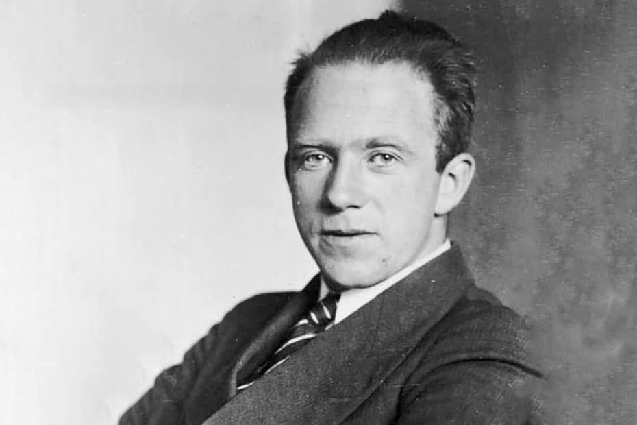 Werner Heisenberg, usando um terno preto e uma gravata listrada preta e branca. Ele tem um olhar sério e focado, com olhos intensos que refletem a profundidade de suas descobertas científicas. Sua expressão transmite a confiança de um dos principais pioneiros da mecânica quântica. O fundo é simples e neutro, destacando sua postura refinada e seu estilo clássico. Heisenberg, conhecido por sua formulação do Princípio da Incerteza, é retratado com um ar de autoridade intelectual.