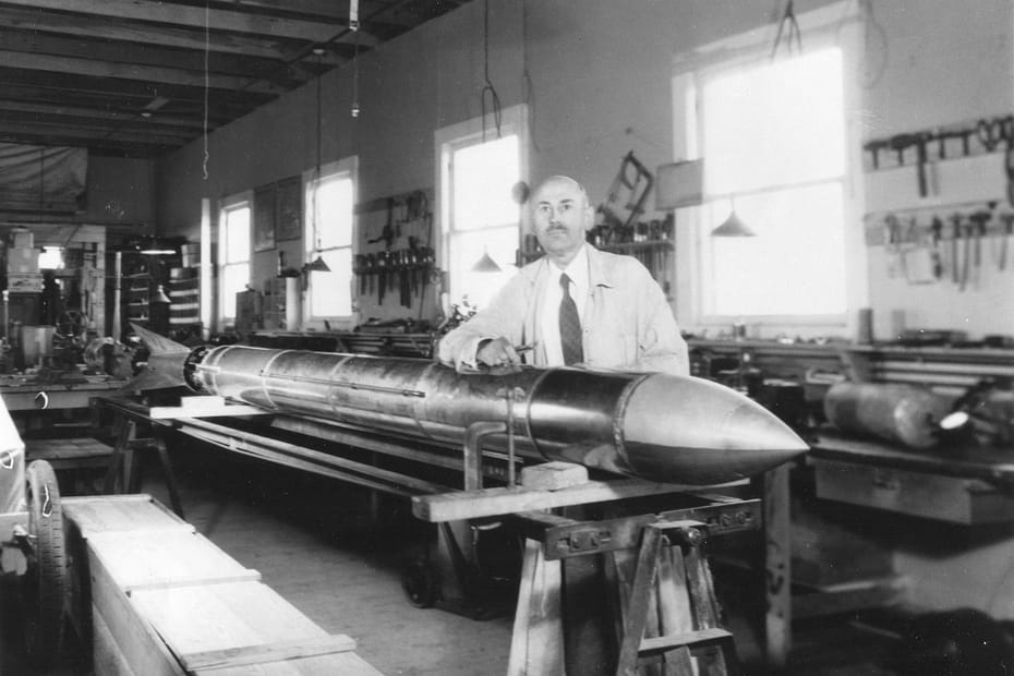 Dr. Robert H. Goddard em frente a um foguete apoiado em uma mesa dentro de um galpão, usando jaleco e gravata. Ao fundo, há ferramentas espalhadas, e o Dr. Goddard tem um olhar sério, focado em seu trabalho