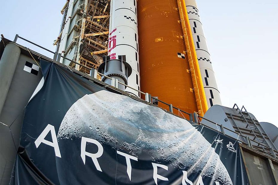 Bandeira do Programa Artemis da NASA em destaque com o imponente foguete SLS ao fundo, representando qual o principal objetivo do Programa Artemis da NASA, que é conduzir missões tripuladas de volta à Lua com tecnologia de ponta.