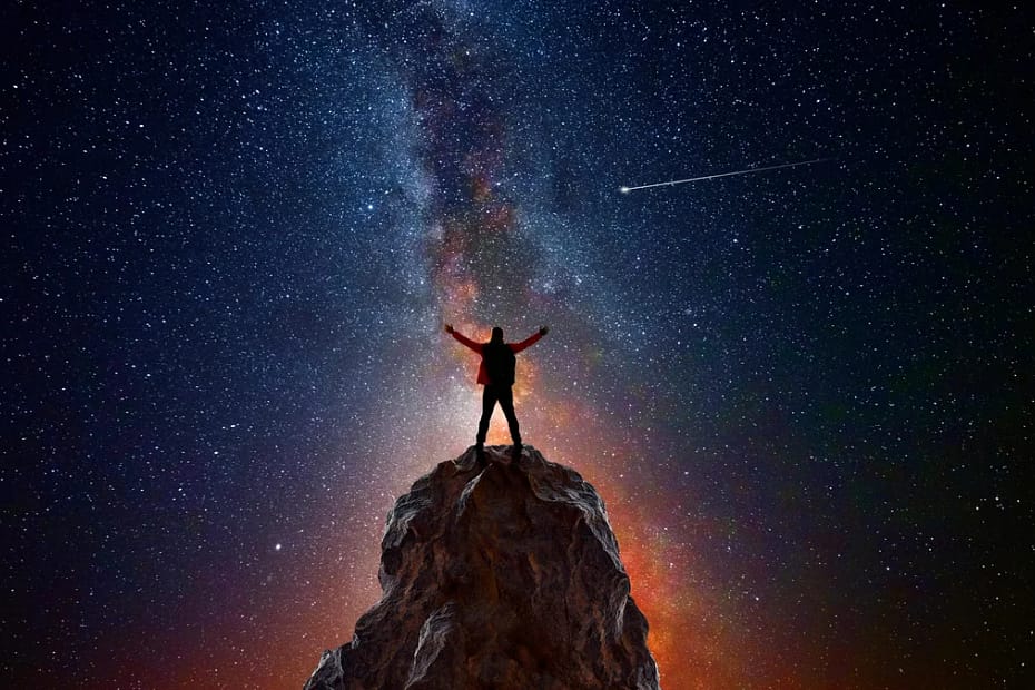 Pessoa no topo de uma montanha com os braços levantados em direção ao céu noturno estrelado, observando um cometa e o braço da Via Láctea, representando como a astronomia define a conexão humana com o universo.