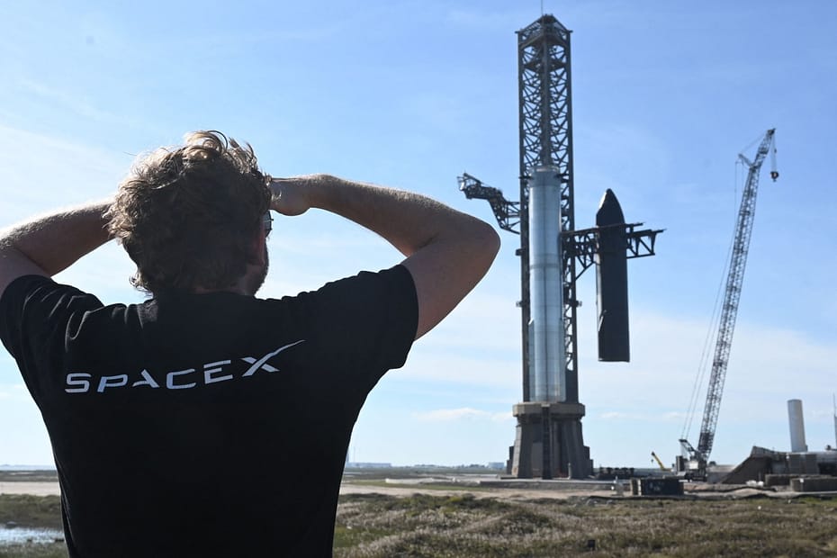 Captura do Super Shuper heavy Elon Musk de costas, vestindo uma camiseta preta com o logo da SpaceX, observa o Super Heavy e a Starship sendo segurados pelos chopsticks do Mechazilla.