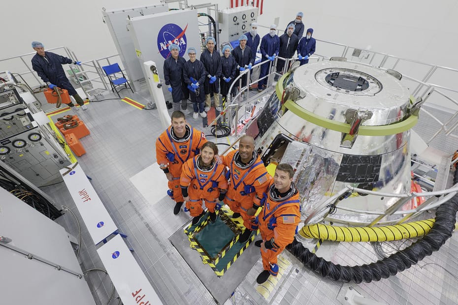 A tripulação da Artemis II (da esquerda para a direita) — Jeremy Hansen, da Agência Espacial Canadense (CSA), especialista de missão; Christina Koch, especialista de missão; Victor Glover, piloto; e Reid Wiseman, comandante — veste seus trajes do Sistema de Sobrevivência da Tripulação Orion para um treinamento de múltiplos dias no módulo da cápsula, iniciado na quinta-feira, 31 de julho de 2025, no Kennedy Space Center, da NASA, na Flórida. Enquanto isso, ao fundo, usando roupas esterilizadas, estão os membros da equipe de encerramento da Artemis II.