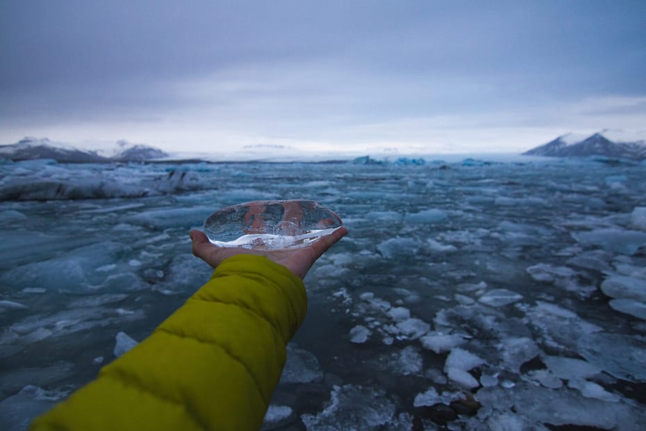 Um braço humano segura um pedaço de gelo diante de uma vasta paisagem de degelo, simbolizando o impacto do aquecimento global.
