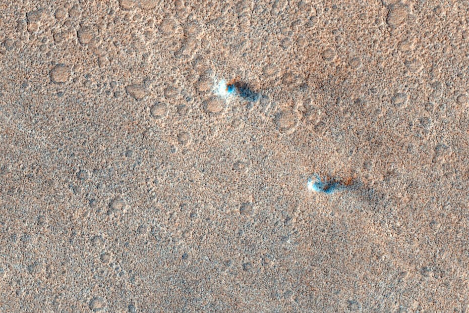 Uma imagem de satélite colorida de Marte mostra vários redemoinhos de poeira — pequenas colunas de poeira em forma de tornados — se movendo sobre uma superfície empoeirada e repleta de crateras. Os redemoinhos aparecem como faixas ou manchas claras, projetando sombras sutis enquanto se deslocam. O terreno apresenta tons avermelhados e amarronzados, com crateras circulares espalhadas e uma textura rugosa.