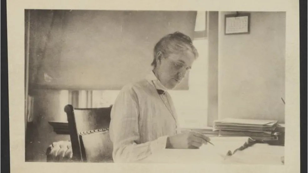 Henrietta Swan Leavitt sentada à mesa, escrevendo em um livro com uma caneta na mão. A imagem representa seu trabalho minucioso na análise das estrelas cefeidas, que revolucionou a medição de distâncias no universo