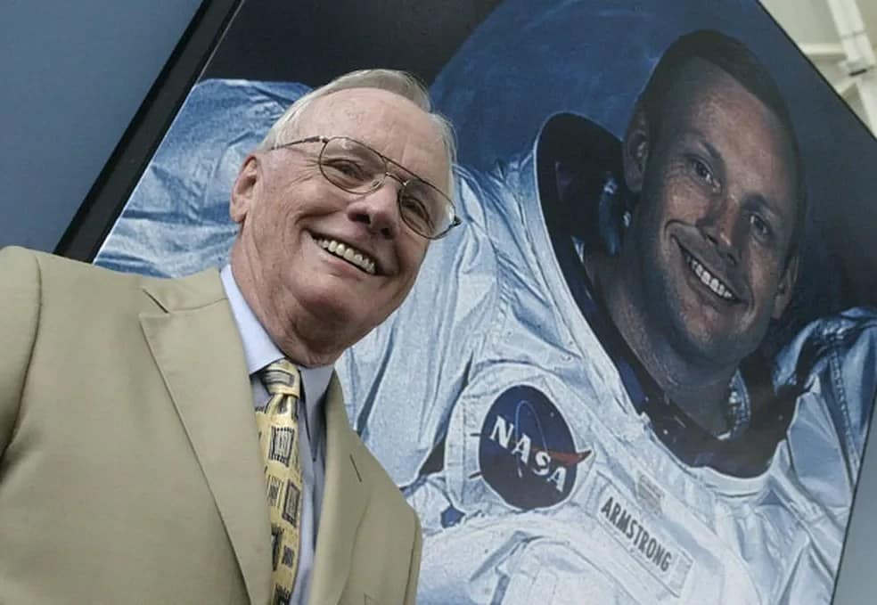 Neil Armstrong, já idoso, sorrindo enquanto está à frente de um banner com sua foto oficial da NASA, onde ele aparece de traje espacial branco. O banner traz a imagem clássica do astronauta  é possível notar lua no fundo de banner ao fundo, simbolizando sua histórica conquista na missão Apollo 11.
