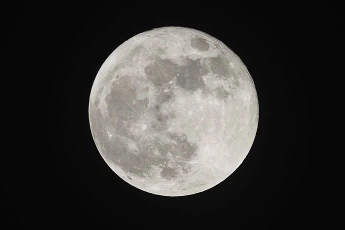 Lua na fase cheia, iluminada de maneira intensa e clara, com detalhes visíveis de suas crateras e mares lunares. O céu ao fundo é de um tom profundo de preto, destacando a beleza e a magnitude da Lua em seu esplendor.
