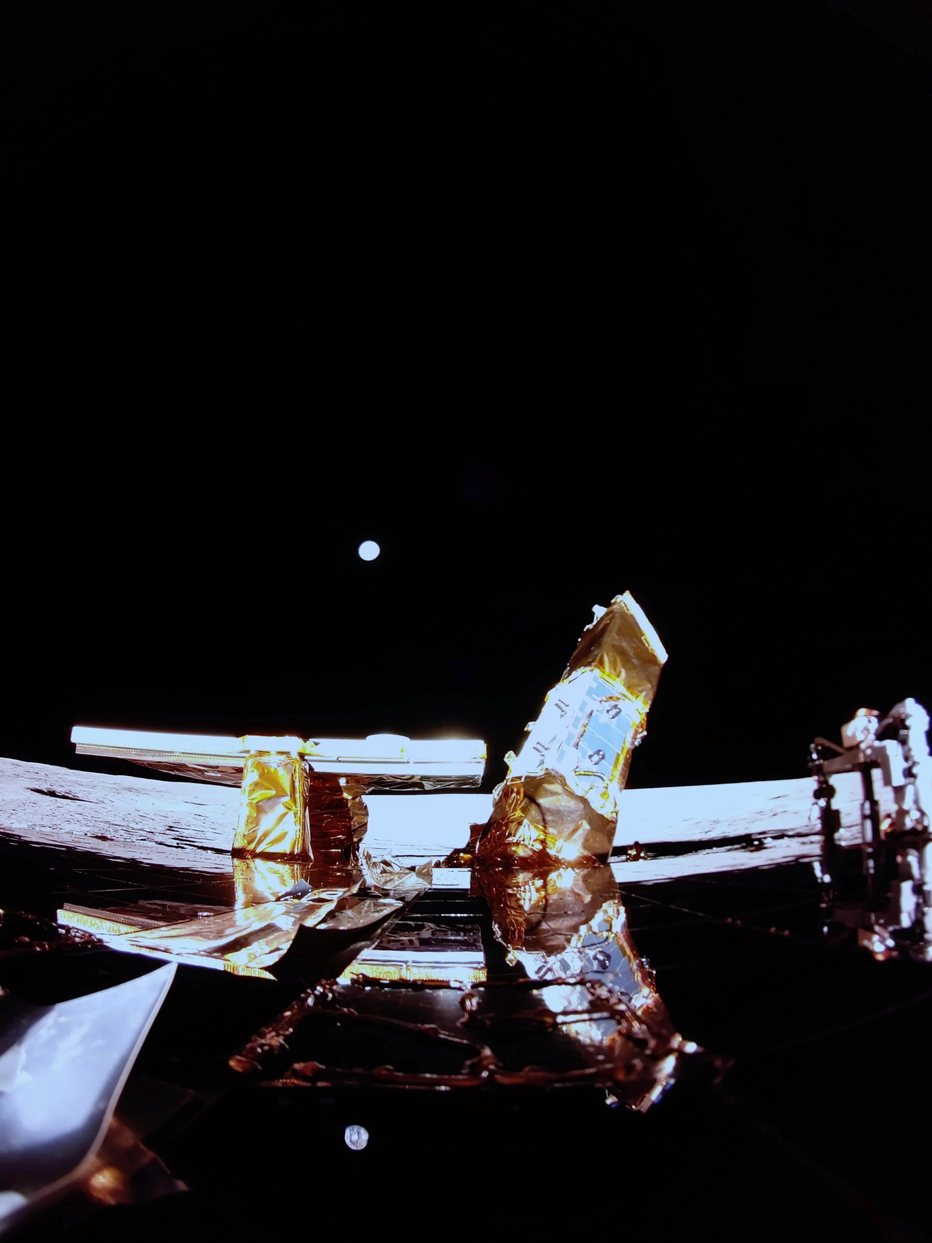 Imagem capturada pelo Blue Ghost, mostrando a superfície lunar com a Terra ao fundo, visível no horizonte. No primeiro plano, é possível ver o convés superior do módulo de pouso, com destaque para o painel solar, a antena de banda X à esquerda e a carga útil LEXI à direita. Esse registro oferece uma visão única da missão, com a Terra distante como pano de fundo, enquanto os equipamentos essenciais para a exploração lunar são claramente visíveis.