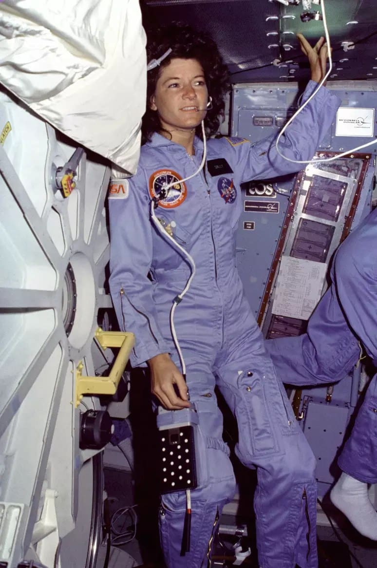 Fotografia histórica de Sally Ride, a primeira mulher americana no espaço, dentro da cabine do ônibus espacial. Ela está com o cabelo volumoso, usando o traje cinza da NASA e fones de ouvido. Ao fundo, é possível ver o painel de controle da nave, destacando a tecnologia da época e a importância da missão espacial por esse feito ela entra na lista de astronautas famosos da nasa