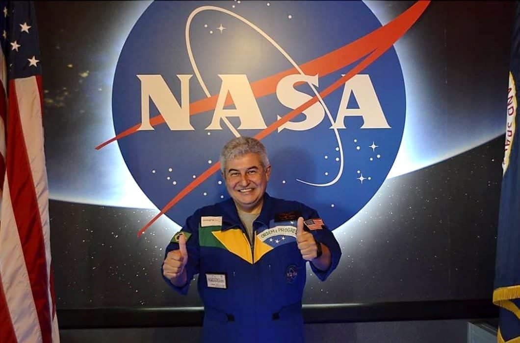 Marcos Pontes, astronauta brasileiro, posa em frente ao logo da NASA. Ele está vestindo um traje espacial azul, com a bandeira do Brasil no peito, e faz o sinal de 'joia' com uma mão