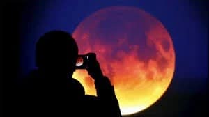 Silhueta de uma pessoa tirando uma foto da Lua de Sangue, com o céu estrelado ao fundo e a Lua brilhando intensamente em tons vermelhos. A imagem captura o momento de admiração e curiosidade, destacando a conexão entre o ser humano e o cosmos