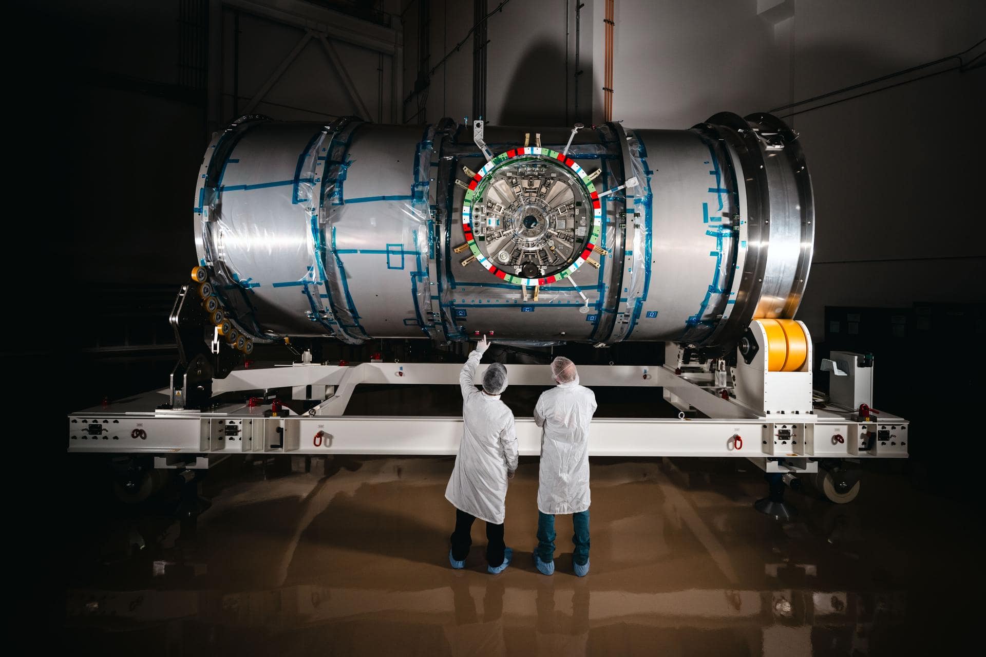 Dois técnicos em trajes de sala limpa estão em frente ao HALO (Habitation and Logistics Outpost) na instalação da Northrop Grumman em Gilbert, Arizona. O módulo cilíndrico metálico, recentemente desembrulhado, está repousando sobre um suporte branco. Um dos técnicos aponta para a parte frontal do módulo. Tecnologias do Programa Artemis da NASA