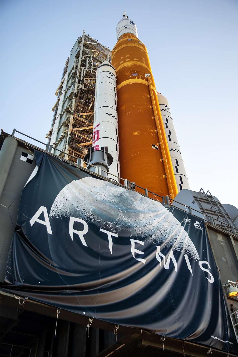 Bandeira do Programa Artemis da NASA em destaque com o imponente foguete SLS ao fundo, representando qual o principal objetivo do Programa Artemis da NASA, que é conduzir missões tripuladas de volta à Lua com tecnologia de ponta.