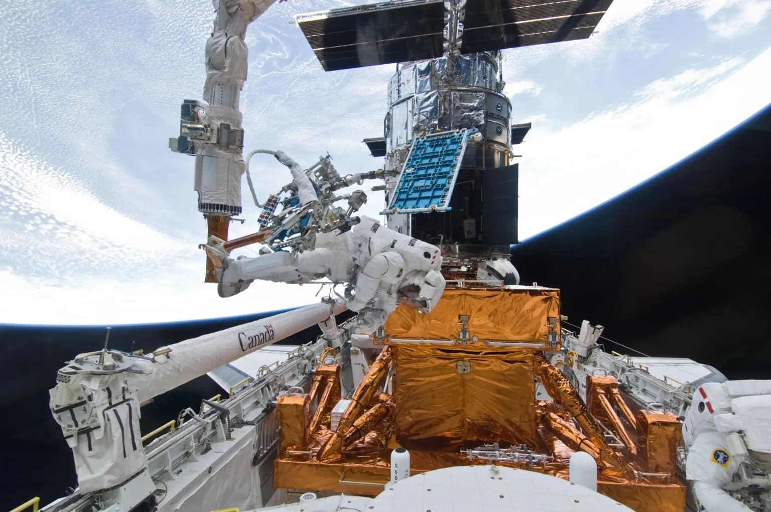 O astronauta Mike Massimino é fotografado do lado de fora da janela do ônibus espacial Atlantis durante uma missão de reparo no Telescópio Espacial Hubble, com a estrutura do telescópio parcialmente visível no fundo.