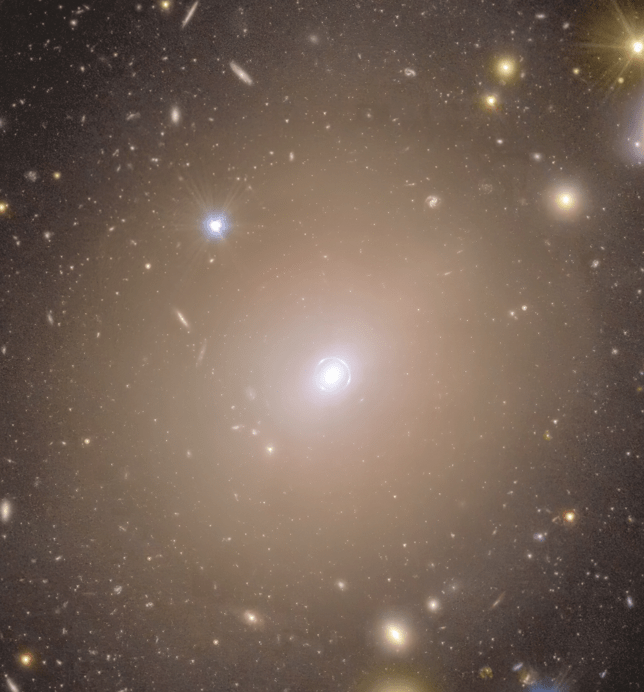 Foco no Anel Um mar de estrelas coloridas e galáxias parece nadar na imensidão escura do espaço ao redor de um halo nebuloso que ocupa o centro da cena. No meio da imagem, o aglomerado difuso de luz, em um tom quente de amarelo, se estende ao redor de um pequeno ponto brilhante, encaixado dentro de um círculo tênue de luz que parece traçado bem de perto ao seu redor. Conforme seguimos a borda do halo central para fora, seu brilho diminui e se mistura suavemente com o ambiente ao redor. Ali, discos alongados em tons que variam de um roxo quente a um dourado amarelado, além de pontos luminosos com pontas de difração bem definidas, se espalham de forma equilibrada por toda a imagem.
