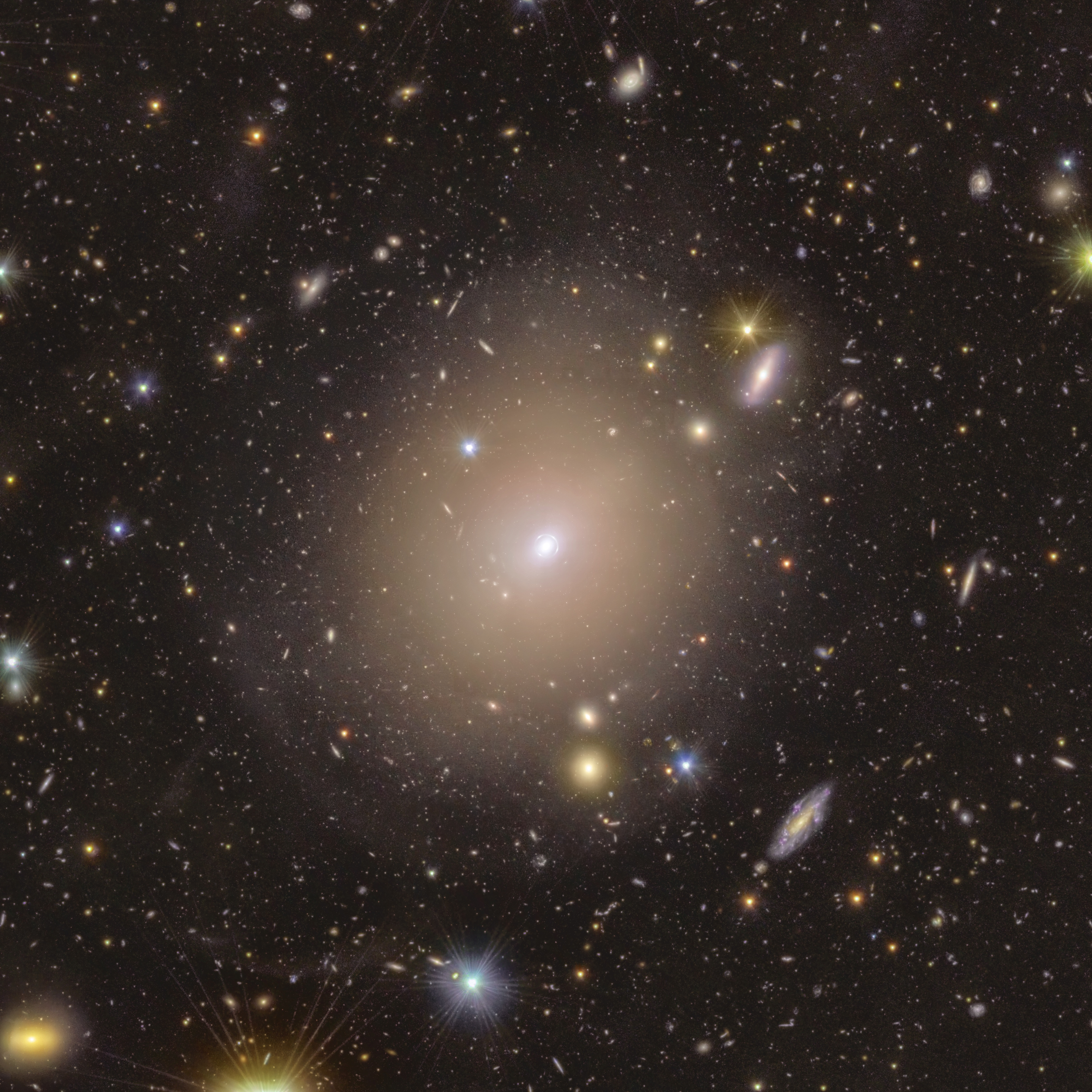 Um mar de estrelas coloridas e galáxias parece nadar na imensidão escura do espaço ao redor de um halo nebuloso que ocupa o centro da cena. No meio da imagem, o aglomerado difuso de luz, em um tom quente de amarelo, se estende ao redor de um pequeno ponto brilhante, encaixado dentro de um círculo tênue de luz que parece traçado bem de perto ao seu redor. Conforme seguimos a borda do halo central para fora, seu brilho diminui e se mistura suavemente com o ambiente ao redor. Ali, discos alongados em tons que variam de um roxo quente a um dourado amarelado, além de pontos luminosos com pontas de difração bem definidas, se espalham de forma equilibrada por toda a imagem.