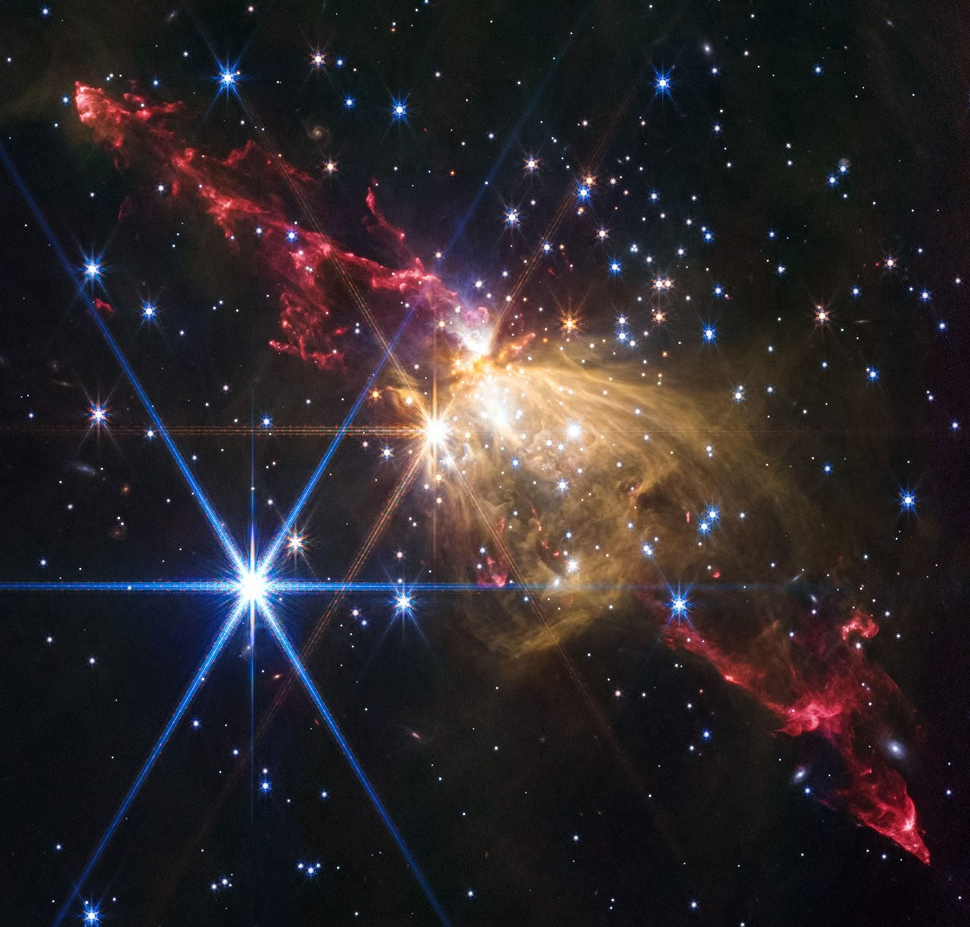  jato estelar gigante Filamentos gasosos amarelo-alaranjados parecem uma rosa vista de lado e inclinada levemente da parte superior esquerda para a inferior direita, um pouco acima do centro da imagem. Estendendo-se da rosa para cima à esquerda e para baixo à direita estão fluxos gasosos que aparecem como lóbulos vermelhos com formato geral de triângulos altos e estreitos, com pontas arredondadas. Cada triângulo vermelho é formado por linhas onduladas e irregulares. Dezenas de estrelas estão espalhadas pelo campo. Uma estrela branca particularmente brilhante, com oito picos de difração, está localizada no topo da rosa amarela. Outra estrela azul brilhante, com picos de difração ainda mais proeminentes, está à sua parte inferior esquerda. O fundo do espaço é preto