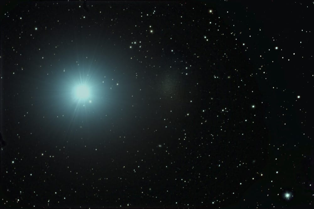 A galáxia anã Leo I aparece como uma mancha tênue e difusa à direita de uma estrela brilhante, destacando-se sutilmente contra o fundo escuro do espaço.