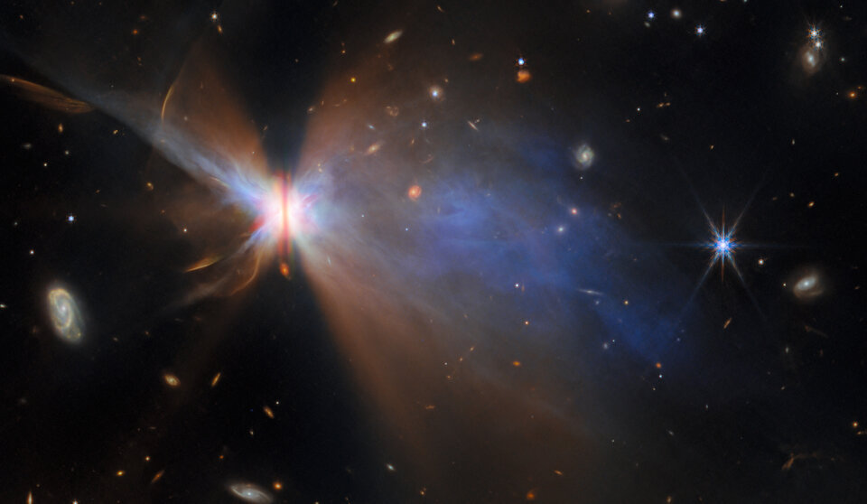 Descrição da imagem: Uma imagem de campo amplo de IRAS 16594-4656 capturada pelo Telescópio Espacial James Webb. O núcleo brilhante da nebulosa é dividido por uma estreita faixa escura, com amplos lóbulos coloridos e luminosos irradiando-se para fora como um arco-íris. Diversas galáxias e estrelas de fundo são visíveis por todo o campo.