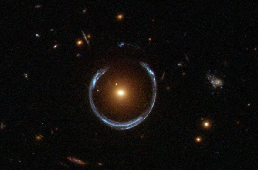 Lente gravitacional da Ferradura Cósmica. O buraco negro ultramassivo recém-descoberto está localizado no centro da galáxia laranja. Bem atrás dela, há uma galáxia azul que está sendo deformada em um anel com formato de ferradura devido às distorções no espaço-tempo causadas pela imensa massa da galáxia laranja em primeiro plano.

Crédito: NASA/ESA
