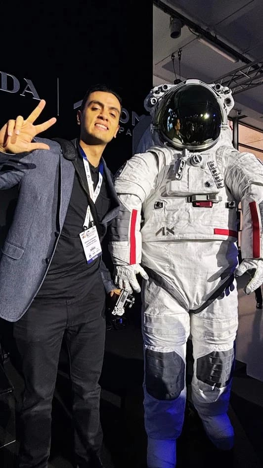 Pessoa vestindo um terno cinza e preto ao lado do traje de exploração espacial da Axion Space, que é branco com capacete e viseira espelhada. O traje apresenta detalhes cinzas nos joelhos e cotovelos, faixas vermelhas nos braços e o logo da Axion na cintura. A foto foi tirada durante o IAC (International Astronautical Congress) em Milão, Itália.